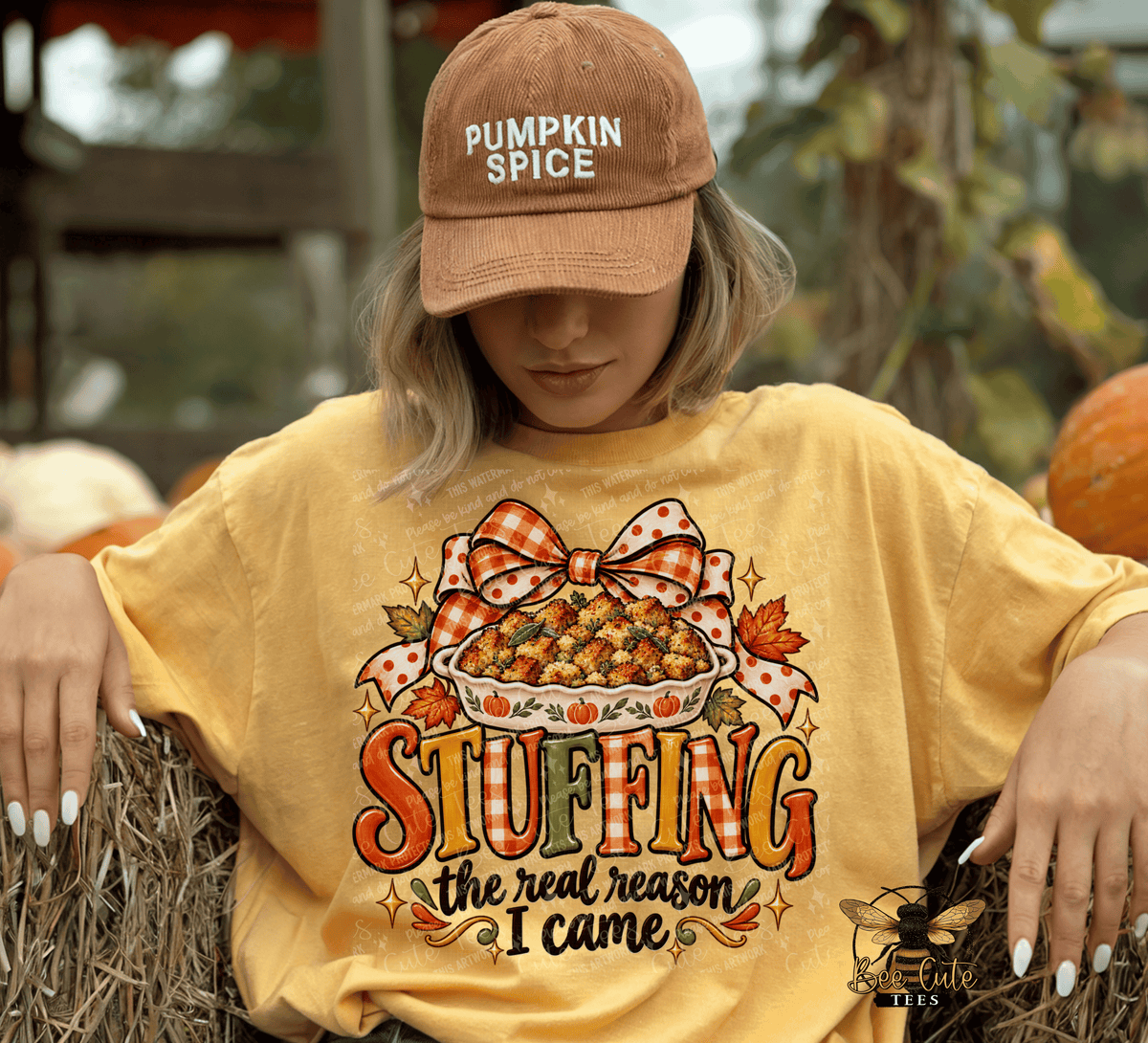 A woman wearing a yellow graphic t-shirt and a brown 'Pumpkin Spice' baseball cap sits on hay bales outdoors. The t-shirt features a graphic of a stuffed pie with a polka-dot bow and the text 'STUFFING the real reason I came'.