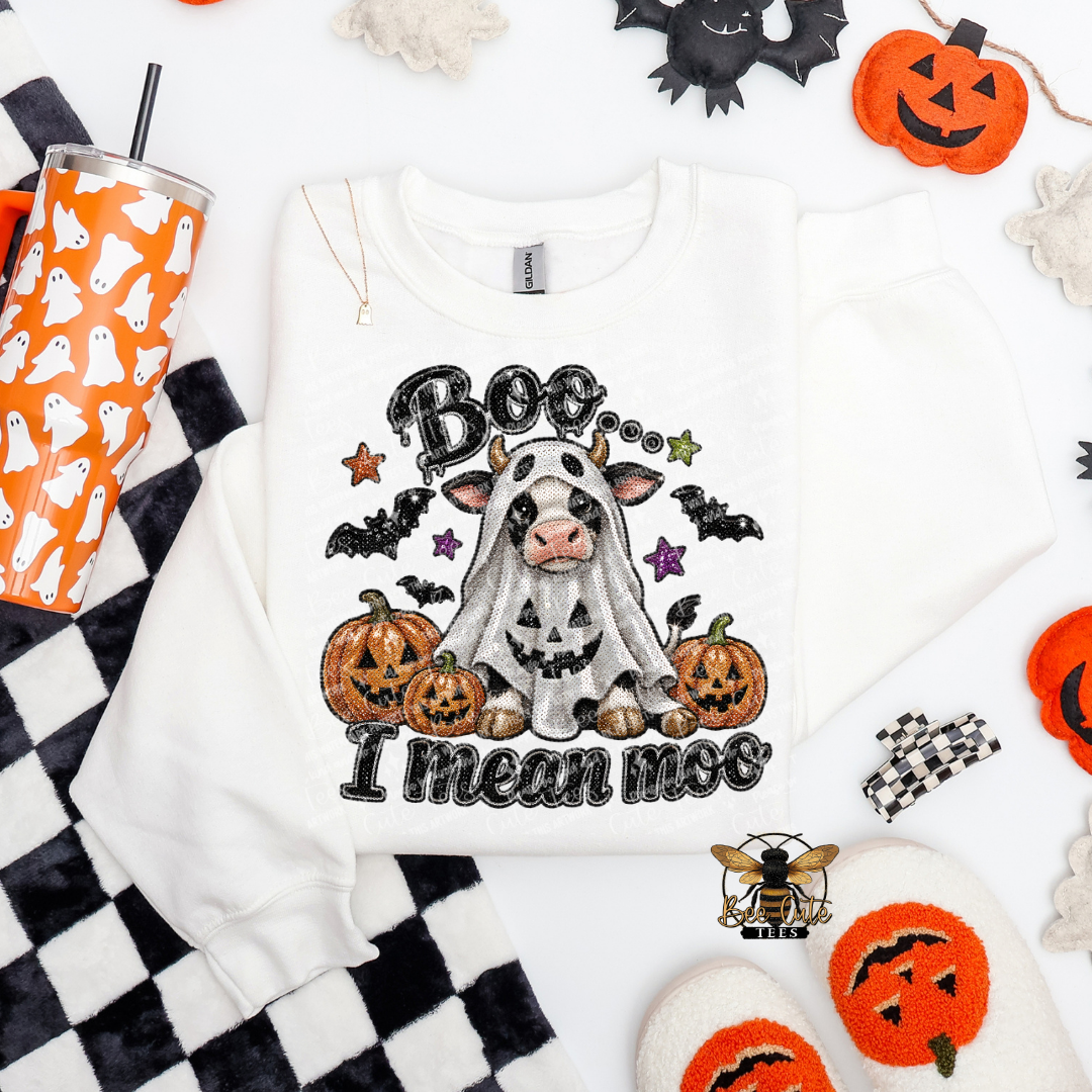 A white crewneck sweatshirt with a Halloween graphic of a cow in a ghost costume surrounded by pumpkins, bats, and the text 'Boo I Mean Moo', styled in a flat lay with seasonal props.
