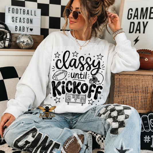 A woman wearing a white crewneck sweatshirt with a black graphic that reads 'classy until KICKOFF' and distressed light-wash jeans, sitting in a decorated room with football-themed decor.