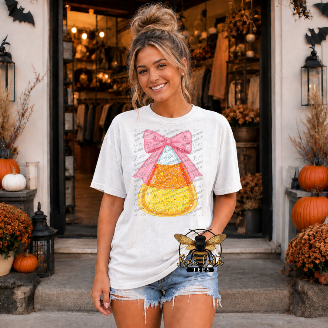 A woman stands in front of a storefront wearing a white graphic T-shirt with a candy corn design and a pink bow, paired with denim shorts. The scene is styled in autumn tones with pumpkins, lanterns, and dried foliage.