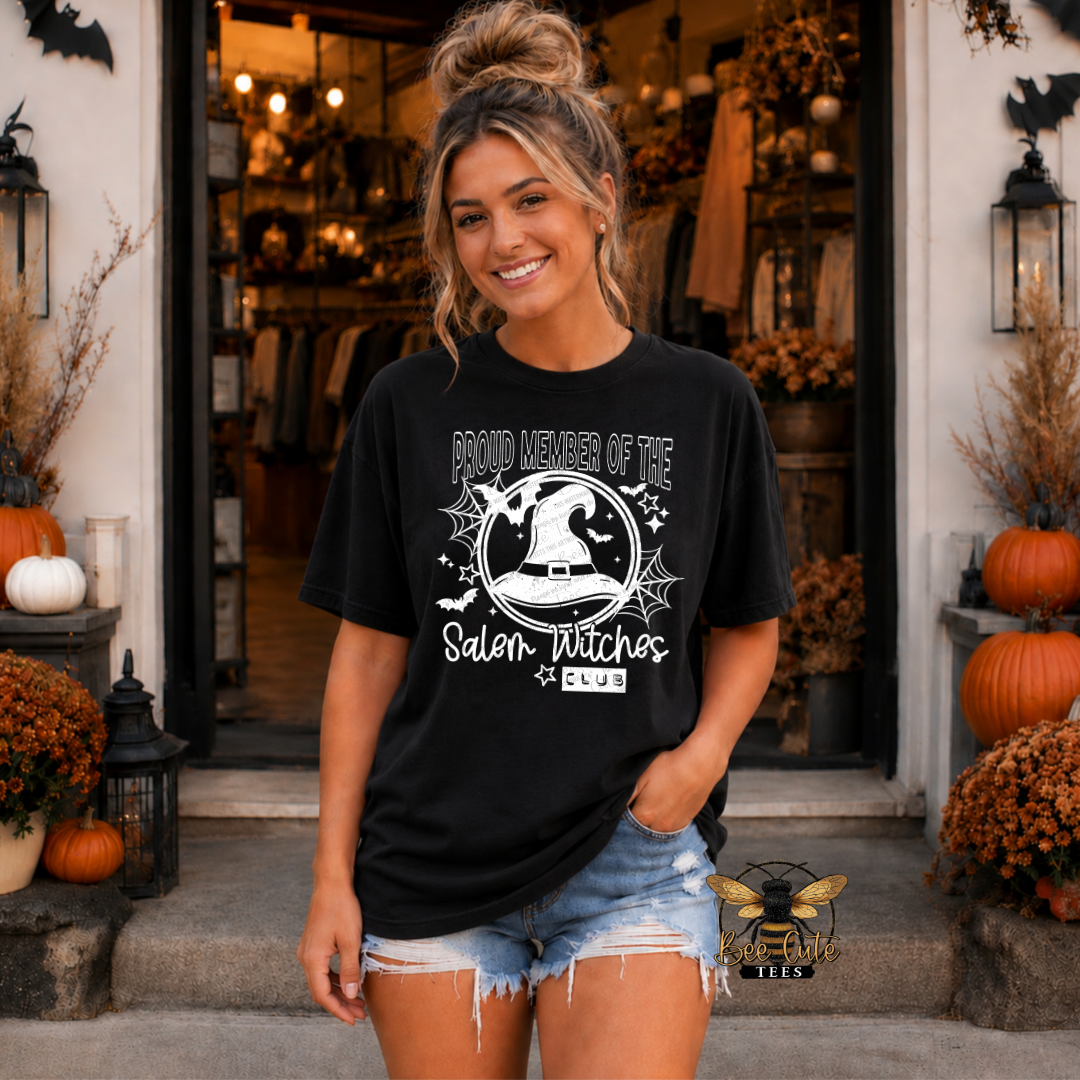 A woman stands in front of a storefront wearing a black graphic T-shirt with a white witch-themed print that reads 'Proud Member of the Salem Witches Club.' She is also wearing light blue denim shorts.