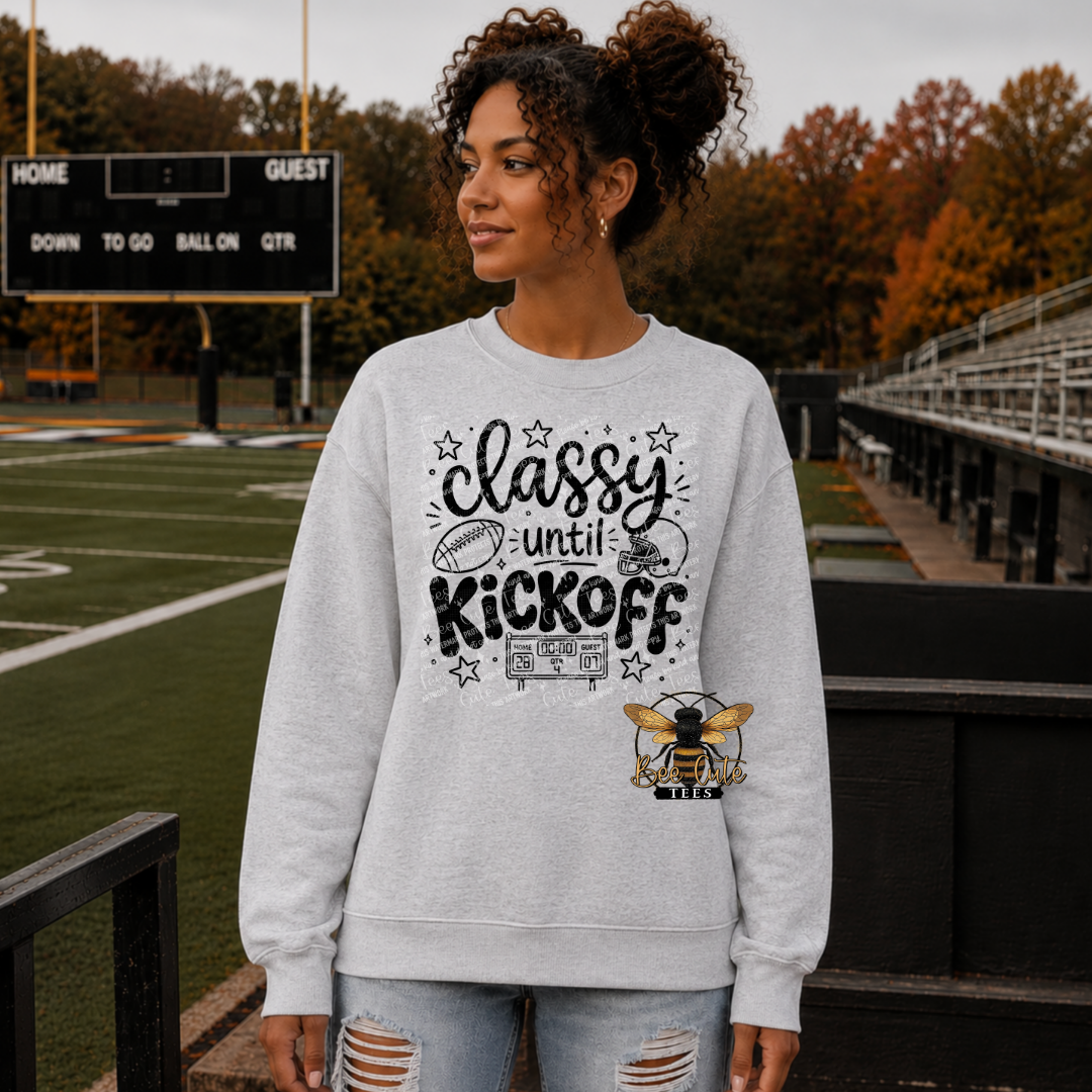 A woman stands on a football field wearing a light gray crewneck sweatshirt with a black graphic that reads 'classy until KICKOFF' and a bee logo, paired with distressed light blue jeans.