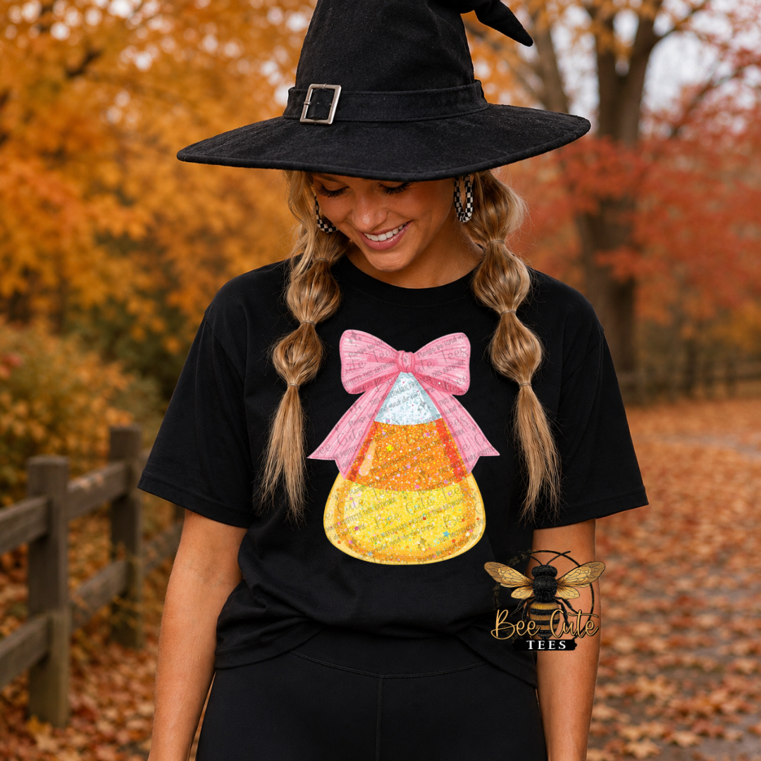 A woman wearing a black witch hat and a black t-shirt with a candy corn graphic featuring a pink bow. She is standing outdoors on a path covered with fallen leaves, with autumn trees in the background.