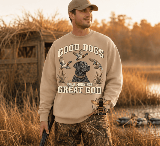 A man wearing a beige crewneck sweatshirt with a graphic print of a black dog and ducks, standing in a marshy field at sunrise. The sweatshirt features the text 'GOOD DOGS COLD MORNINGS GREAT GOD'.