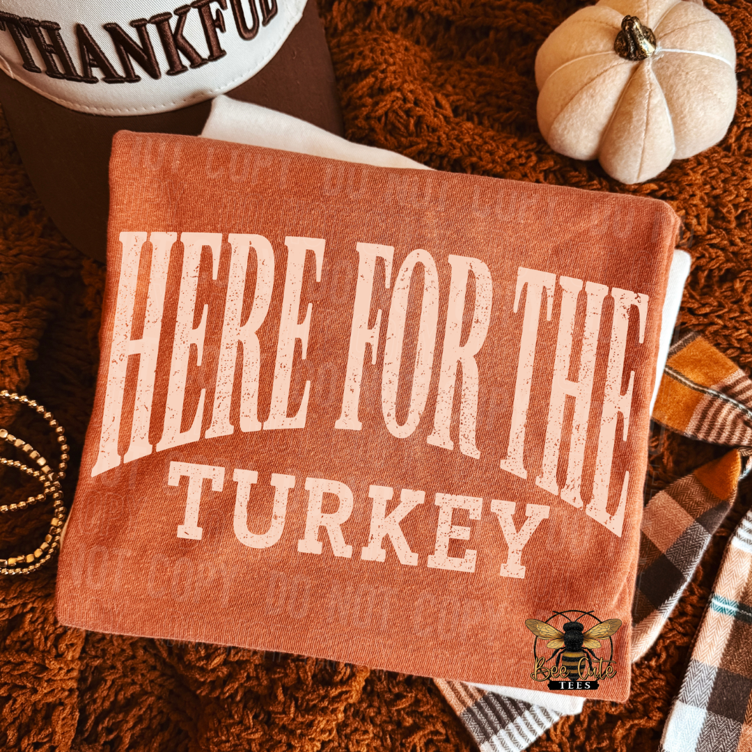 A folded terracotta orange T-shirt with large distressed cream text reading “HERE FOR THE TURKEY,” styled on a brown shag rug with a small pumpkin, plaid scarf, beads, and part of a hat visible around it.