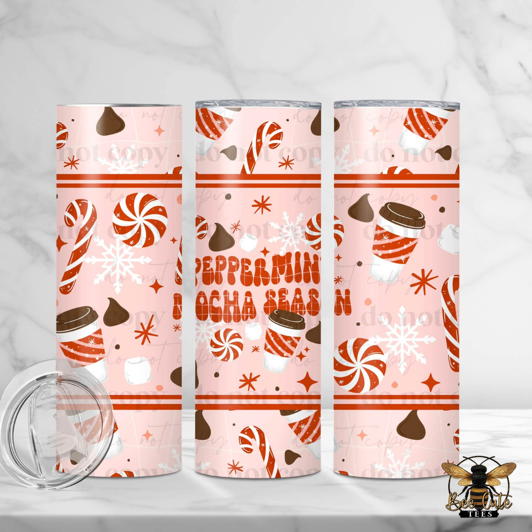 Three tall insulated tumblers with a light pink holiday pattern featuring peppermint candies, chocolate kisses, snowflakes, and the words 'PEPPERMINT MOCHA Season' in orange. A clear lid is shown separately at the lower left on a white marble surface.