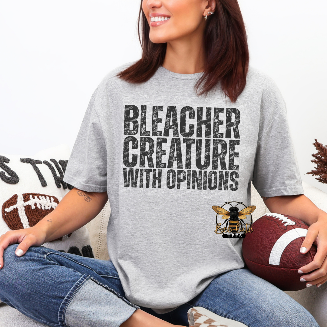 A woman wearing a light gray graphic t-shirt with the text 'BLEACHER CREATURE WITH OPINIONS' and a bee logo, sitting cross-legged and holding a football.
