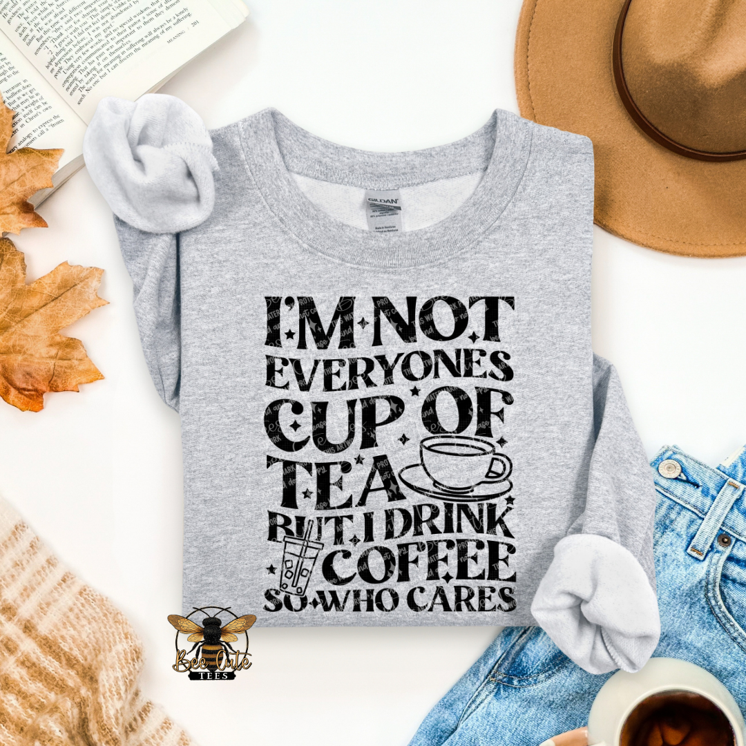 A heather gray long-sleeve t-shirt with a black graphic text that reads 'I'M NOT EVERYONE'S CUP OF TEA BUT I DRINK COFFEE SO WHO CARES' with a coffee cup illustration. The shirt is styled in a flat-lay arrangement with a tan hat, blue jeans, a book, autumn leaves, a cup of coffee, and a knit sweater on a white background.