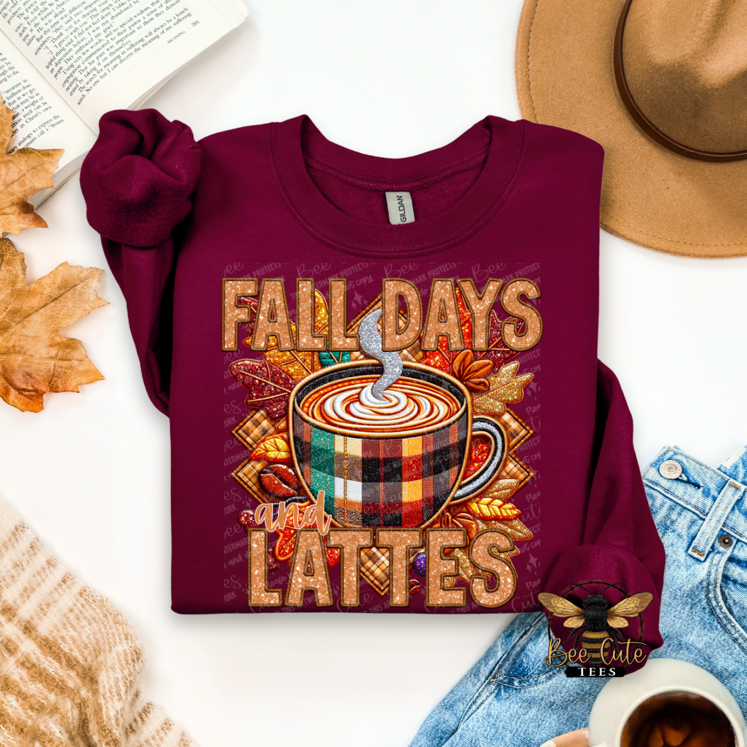 A folded burgundy long-sleeve t-shirt with a 'Fall Days Lattes' graphic, styled in an autumn flat-lay with a tan hat, blue jeans, a book, and a knit sweater on a white background.