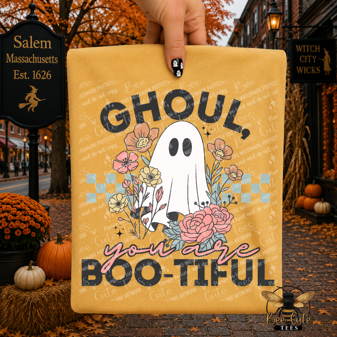 A hand holds a yellow graphic T-shirt featuring a ghost and flowers with the text 'GHOUL, you are BOO-TIFUL' in a Halloween-themed outdoor scene with pumpkins and autumn street decor.