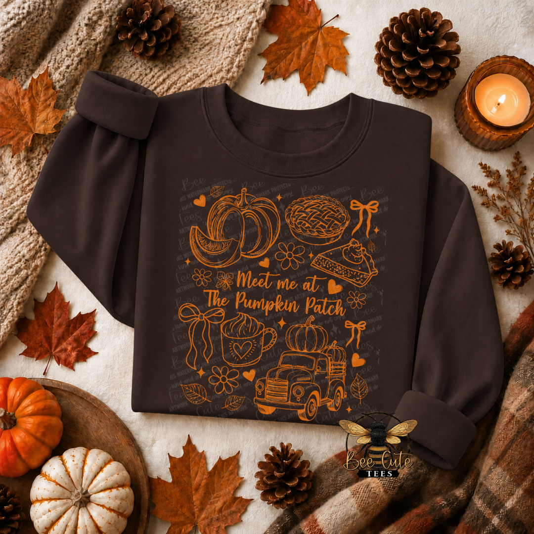 A dark brown long-sleeve sweatshirt with a fall-themed graphic print reading 'Meet me at The Pumpkin Patch' and illustrations of pumpkins, a pie, a truck, and autumn motifs. The sweatshirt is styled in a cozy flat-lay arrangement with pinecones, autumn leaves, a knitted blanket, a small pumpkin, a candle, and a plaid scarf.