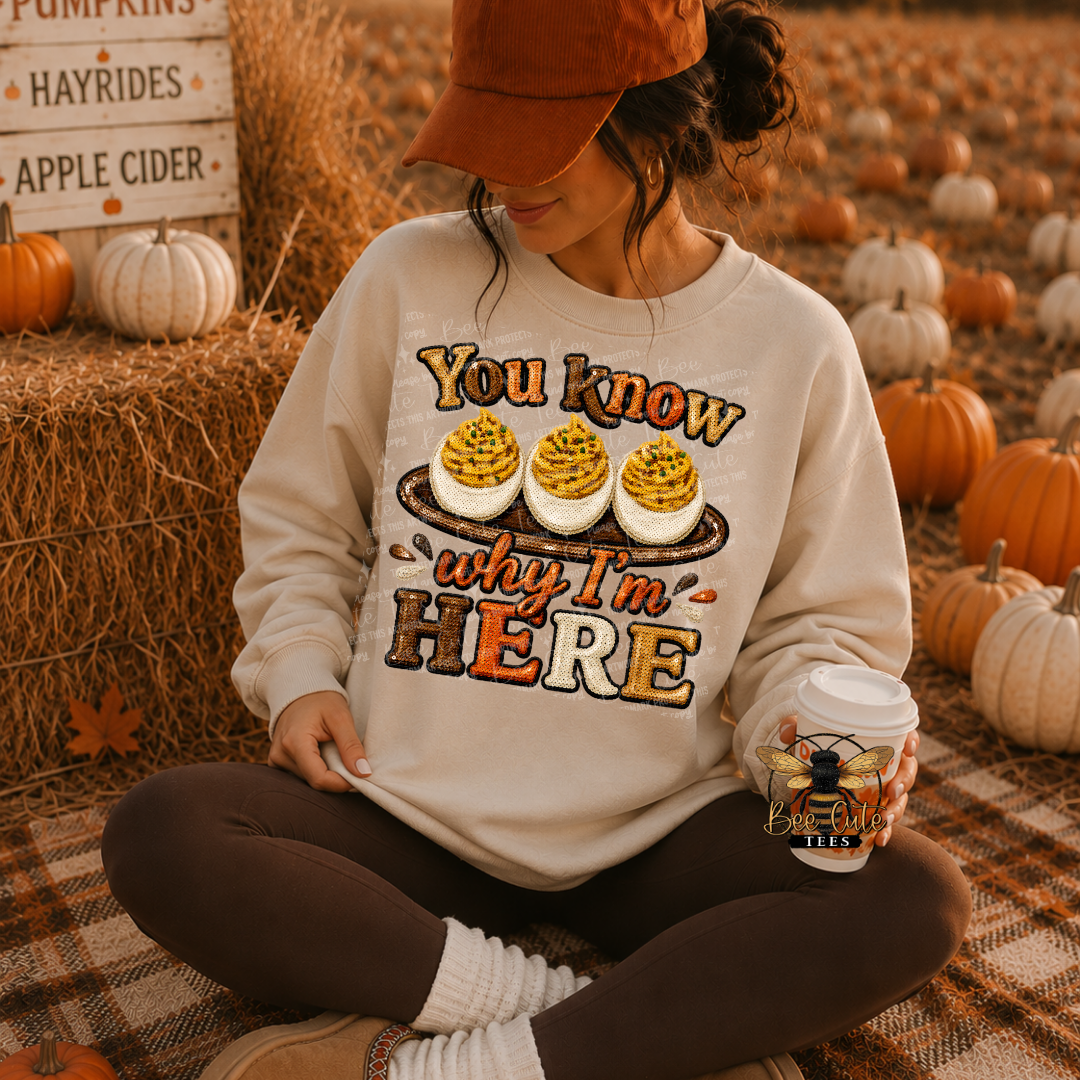 A woman sitting cross-legged in a pumpkin patch wearing a cream sweatshirt with a pumpkin pie graphic and the text 'You know why I'm HERE'. She is wearing a rust-colored cap, dark brown leggings, and holding a coffee cup.