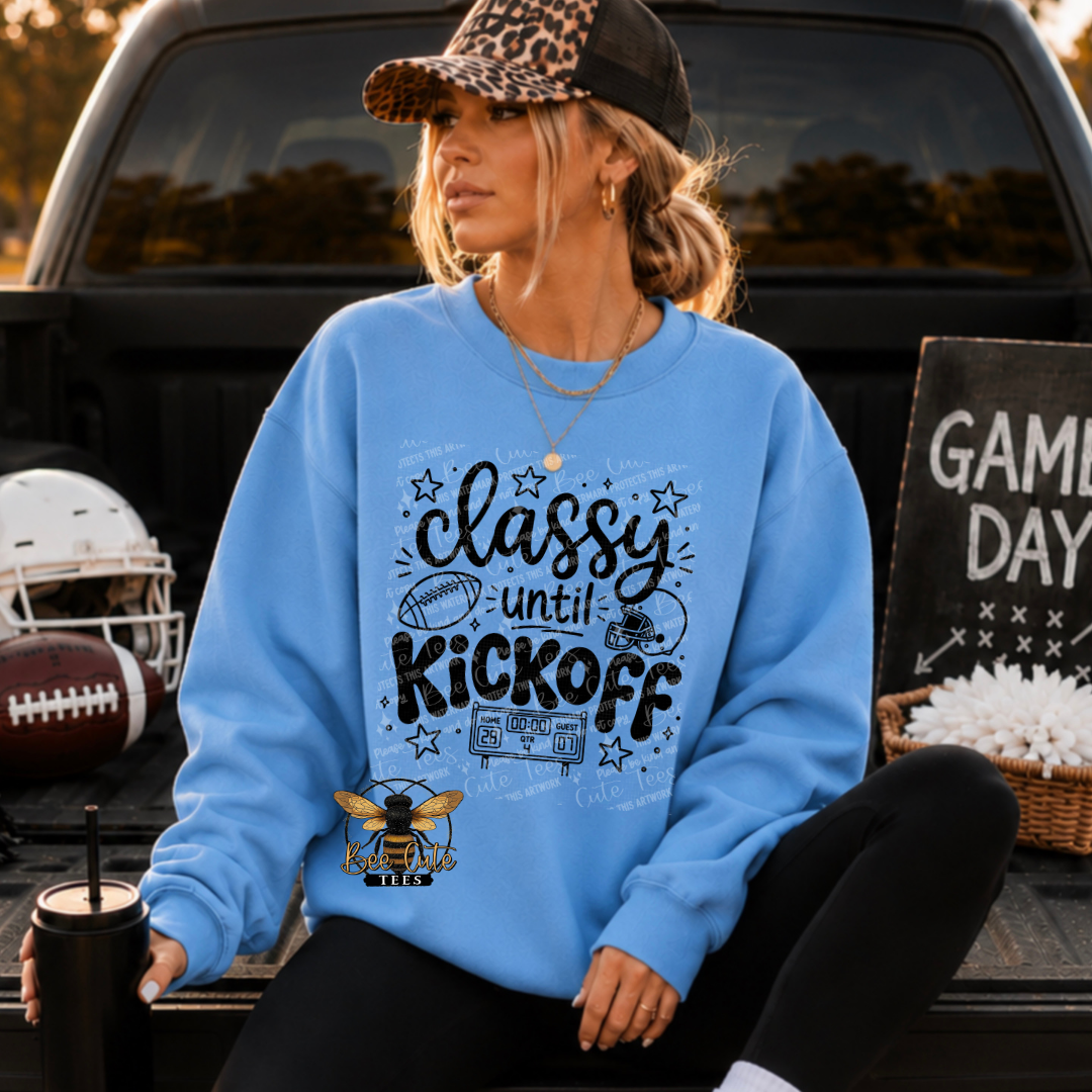 A woman sitting in the bed of a pickup truck wearing a light blue crewneck sweatshirt with a 'classy until KICKOFF' graphic. The scene is styled with football props, including a helmet, a football, a black tumbler, and a 'GAME DAY' sign.