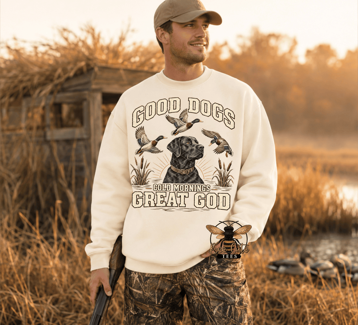 A man wearing a cream-colored crewneck sweatshirt with a graphic of a black labrador retriever and ducks, standing in a marshy outdoor setting at sunrise.