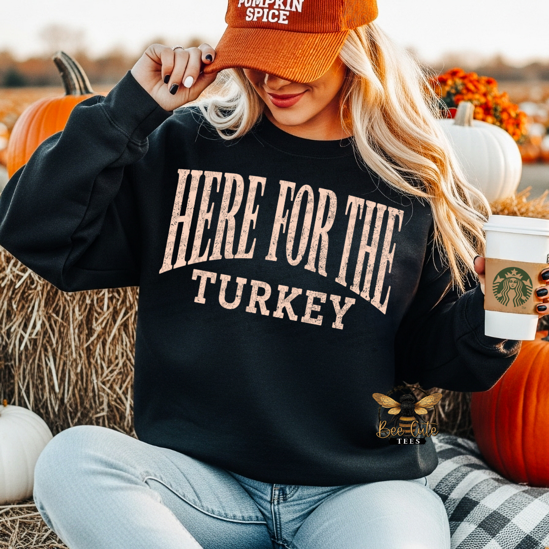 A woman sitting outdoors among pumpkins and hay bales wears a black sweatshirt with the phrase “HERE FOR THE TURKEY” in large beige letters. She also wears an orange cap and holds a coffee cup.