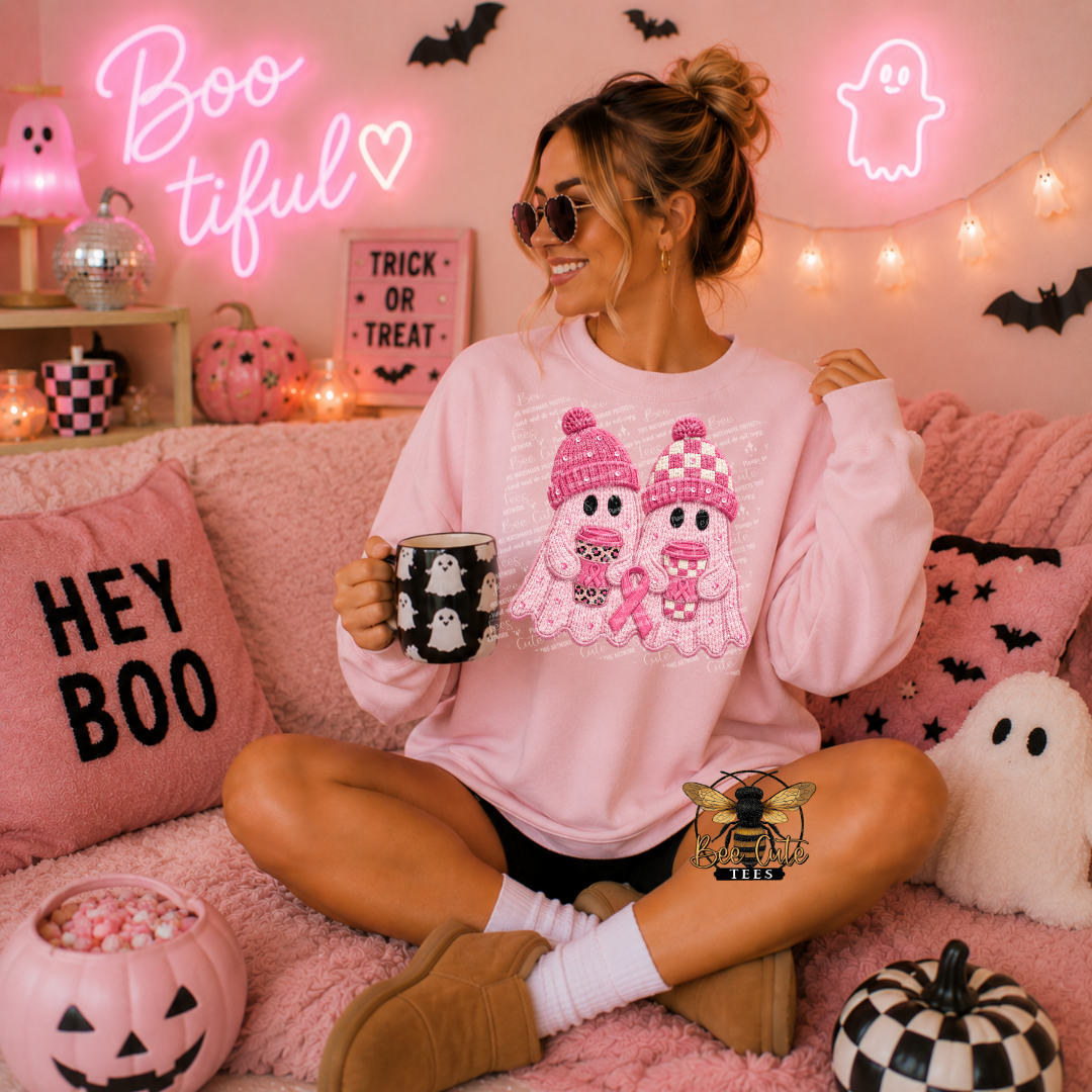 A woman sits cross-legged on a pink couch wearing a light pink sweatshirt with a cute ghost graphic, holding a mug with a ghost pattern. The background is decorated for Halloween with pink pillows, a 'HEY BOO' pillow, a jack-o'-lantern bowl, and glowing pink neon signs.