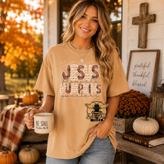 A woman modeling a tan graphic t-shirt with 'JESUS Flannels Pumpkins SWEATERS' in a distressed pattern, holding a white mug with 'BE STILL' on a rustic porch decorated with pumpkins and autumn decor.