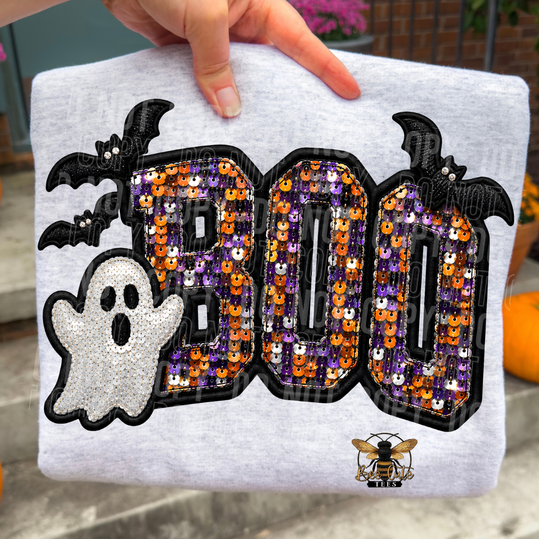 A folded light heather gray T-shirt is held up by a hand, showing a large Halloween graphic on the front. The design features the word 'BOO' in sequined letters with black outlines, a white ghost with black and purple sequins, and two black bats.