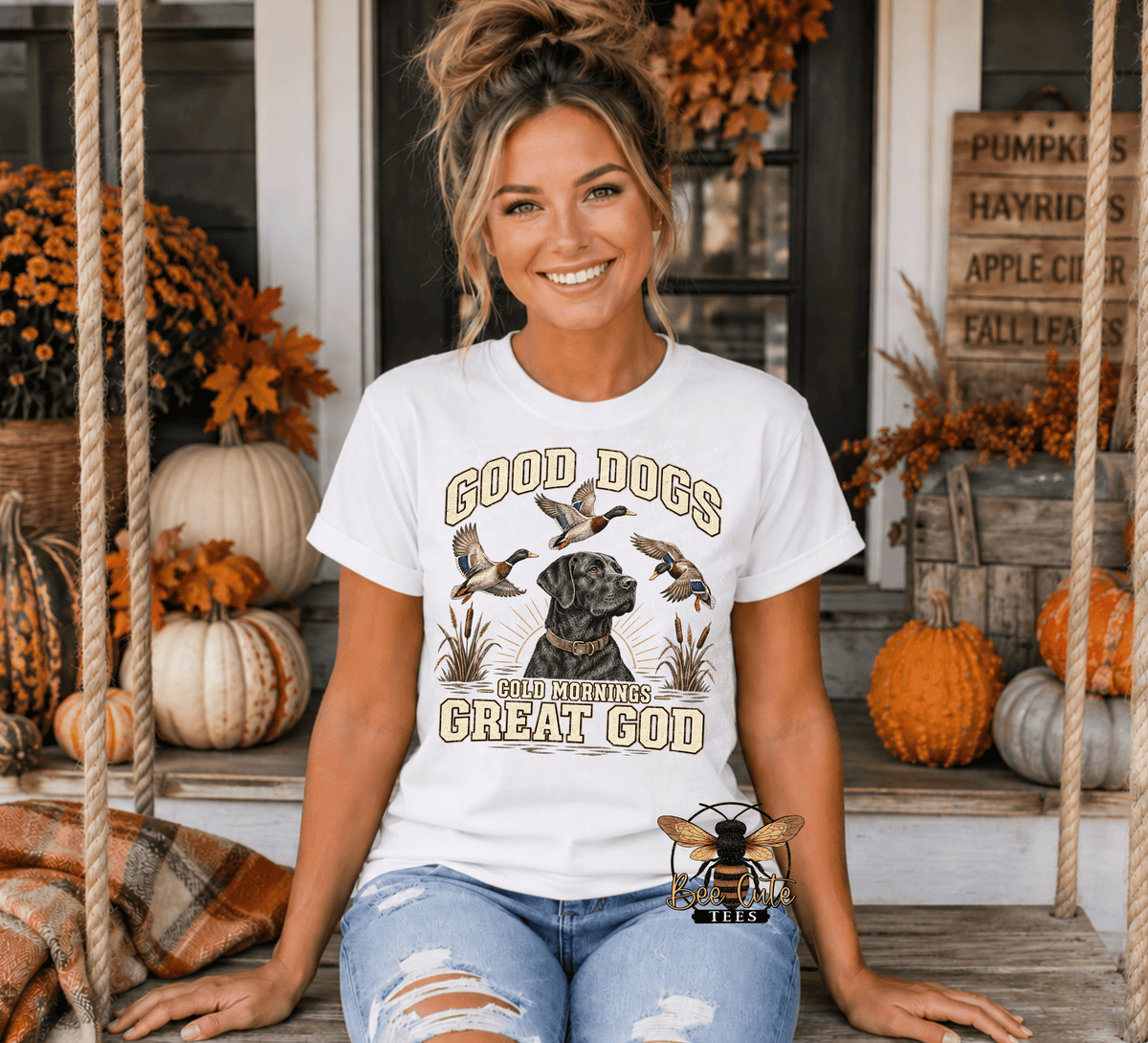 A woman sitting on a porch swing wearing a white graphic t-shirt that reads 'GOOD DOGS COLD Mornings GREAT GOD' with a dog and ducks illustration. The background shows autumn decorations including pumpkins, fall leaves, and rustic wooden signs.