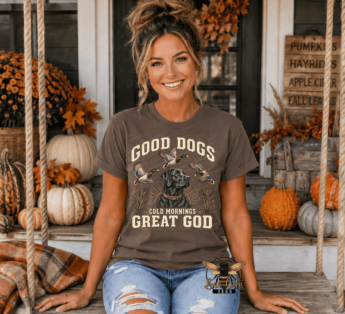 A woman wearing a brown graphic t-shirt with the text 'GOOD DOGS COLD MORNINGS GREAT GOD' and a dog illustration, posing on a rustic wooden porch swing surrounded by autumn pumpkins and fall decorations.