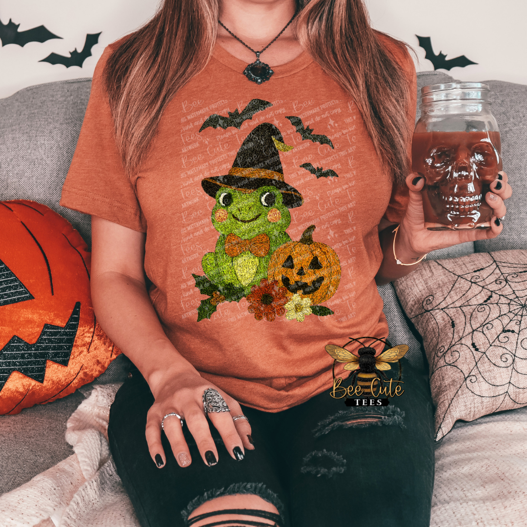 A woman sitting on a couch wearing a rust-orange graphic t-shirt with a green frog and jack-o'-lantern design, holding a skull-shaped glass mug. The shirt features a 'Bee-Cute Tees' brand watermark overlay.