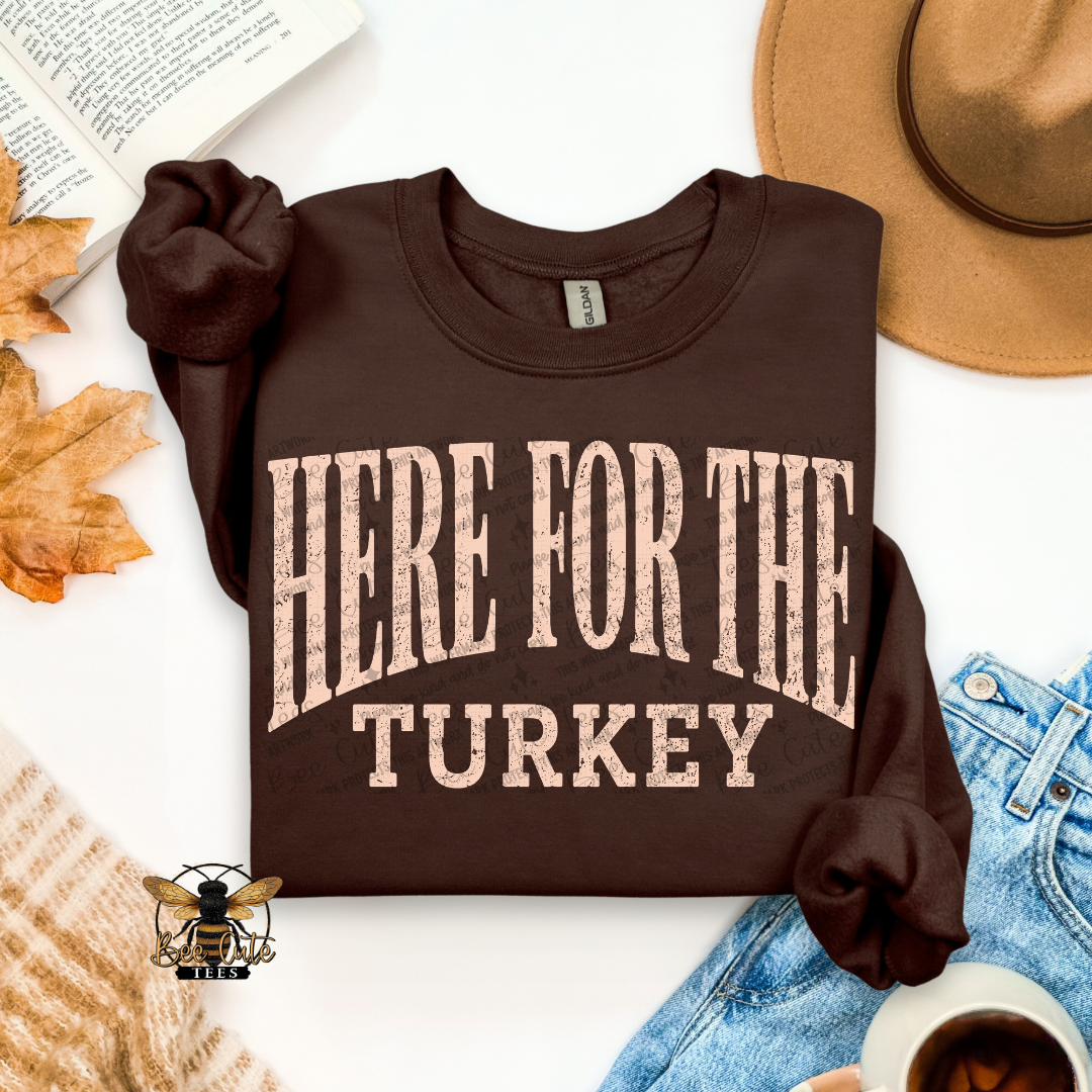 A folded dark brown long-sleeve T-shirt with rolled cuffs, featuring the phrase 'HERE FOR THE TURKEY' in distressed beige lettering. The shirt is styled in a flat lay with autumn leaves, an open book, a tan hat, blue jeans, a cup of coffee, and a knit sweater on a white background.