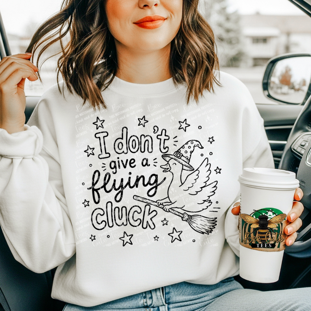 A woman in a car wearing a white sweatshirt with a black graphic of a witch's broom, a flying bird, and the text 'I don't give a flying cluck'. She is holding a white coffee cup with a green and gold logo.