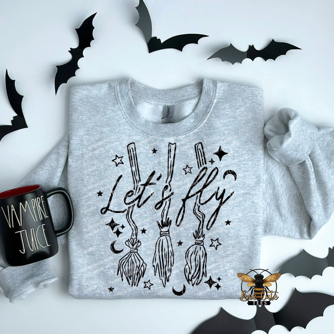 A light heather gray sweatshirt laid flat on a white background, featuring a black graphic of three broomsticks with stars and crescent moons and the phrase 'Let's fly.' A black mug with 'Vampire Juice' is partly visible on the left, and black bat cutouts decorate the scene.