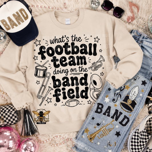 A flat-lay of a beige crewneck sweatshirt with a black graphic text reading 'what's the football team doing on the band field' surrounded by musical instruments and stars. The sweatshirt is styled with light-wash denim jeans decorated with 'BAND' and musical notes, a navy and white baseball cap, sunglasses, and checkered shoes on a textured cream rug.