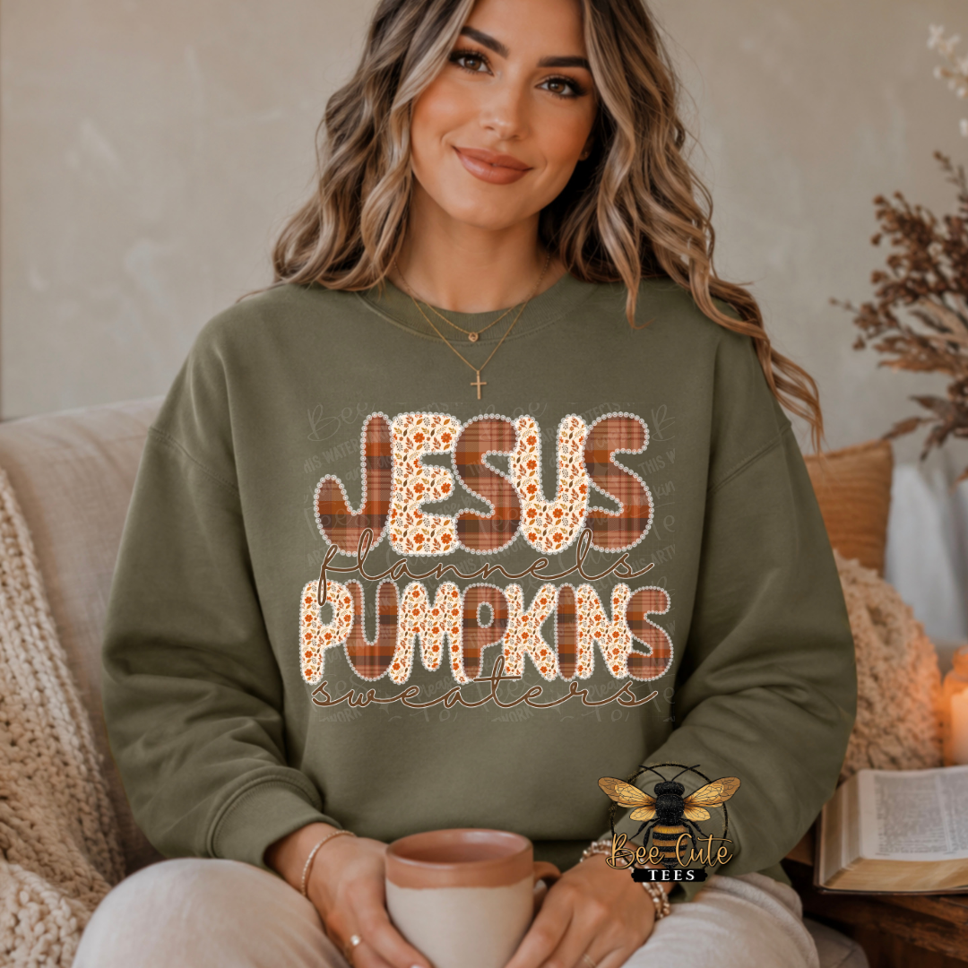 A woman wearing an olive green sweatshirt with a 'JESUS PUMPKINS' graphic sits on a beige couch holding a mug. The graphic features 'JESUS' in a plaid pattern and 'PUMPKINS' in a pumpkin print.