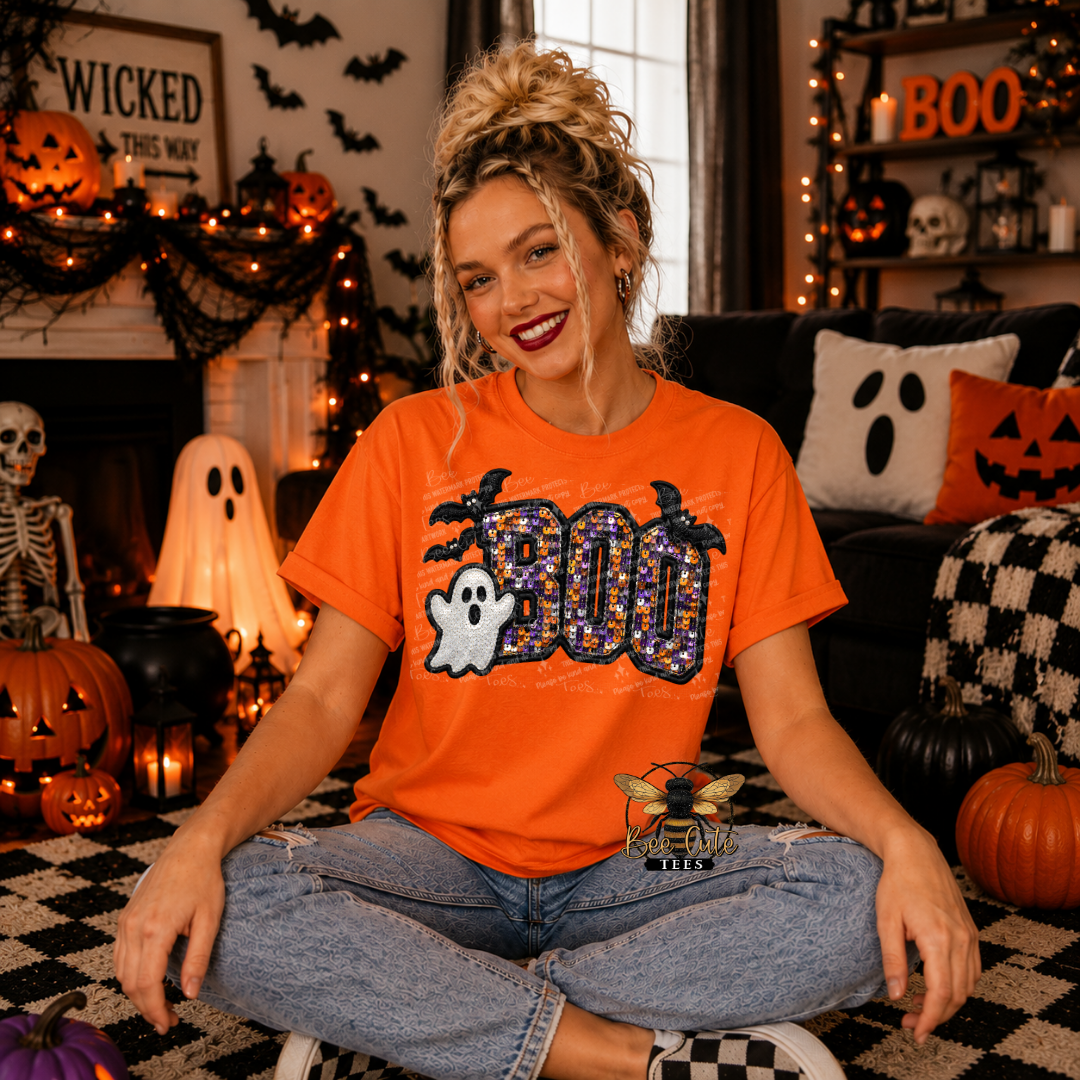 A woman sitting cross-legged in a cozy, decorated Halloween living room, wearing an orange graphic t-shirt with a 'BOO' design featuring a ghost and bats.
