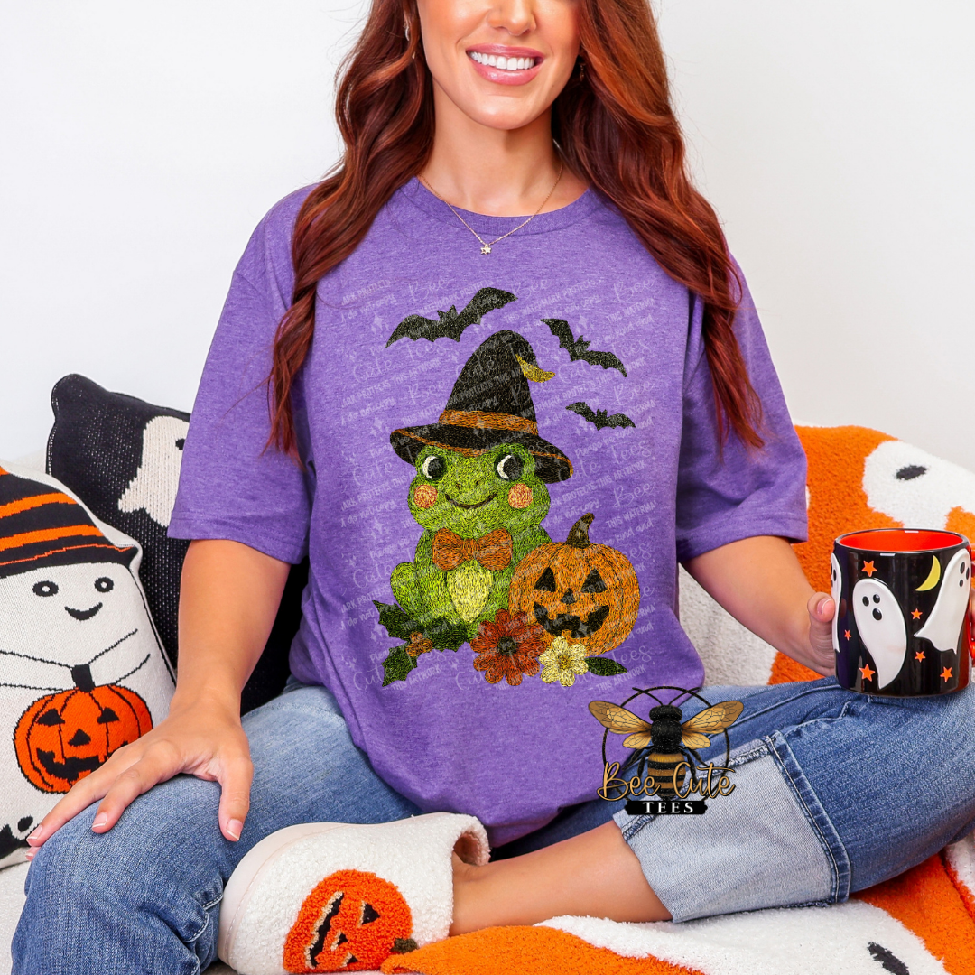 A woman wearing a purple short-sleeve t-shirt with a green cartoon frog wearing a witch hat and surrounded by pumpkins, bats, and flowers. She is sitting on an orange and white blanket with Halloween-themed pillows and slippers, holding a black mug with a ghost design.