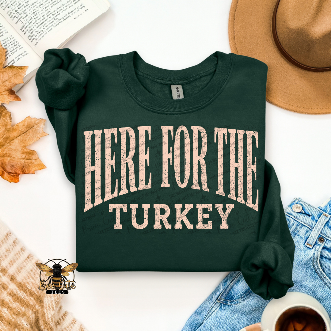 A dark forest green long-sleeve t-shirt with 'HERE FOR THE TURKEY' printed in distressed beige lettering, styled in a flat-lay with a tan hat, blue jeans, a book, autumn leaves, a cup of coffee, and a knit sweater on a white background.