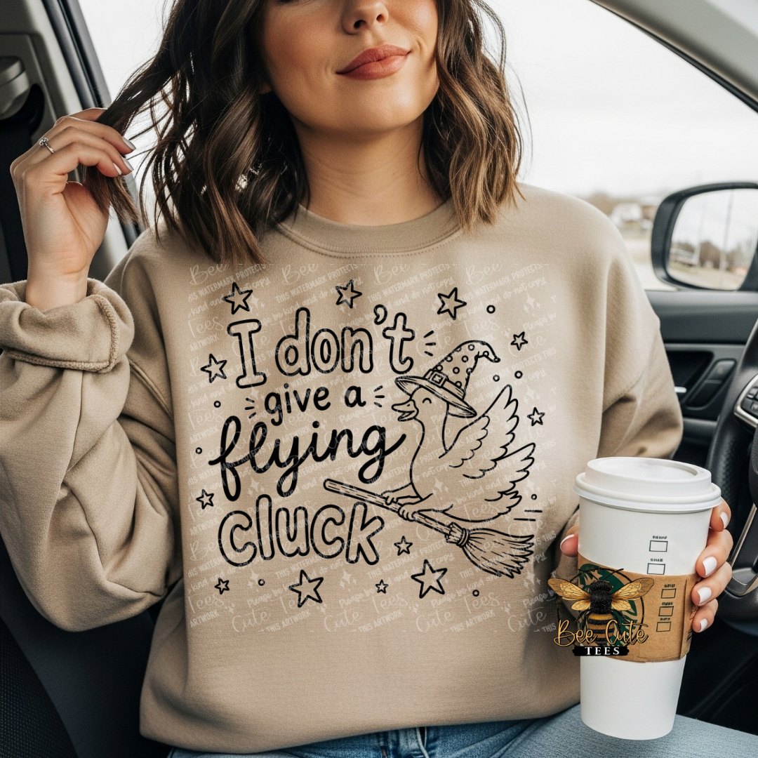 A woman sitting in a car wearing a beige crewneck sweatshirt with a black graphic of a witch, broom, and the text 'I don't give a flying cluck'. She is holding a white coffee cup with a brown sleeve.