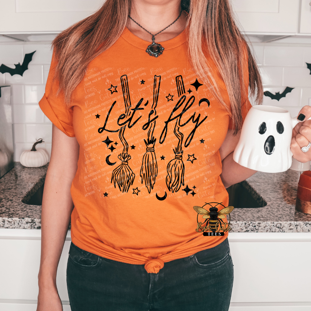 A woman wearing a bright orange graphic t-shirt with a Halloween design featuring broomsticks, stars, moons, and the text 'Let's fly'. She is holding a white ghost-shaped mug in a kitchen setting.