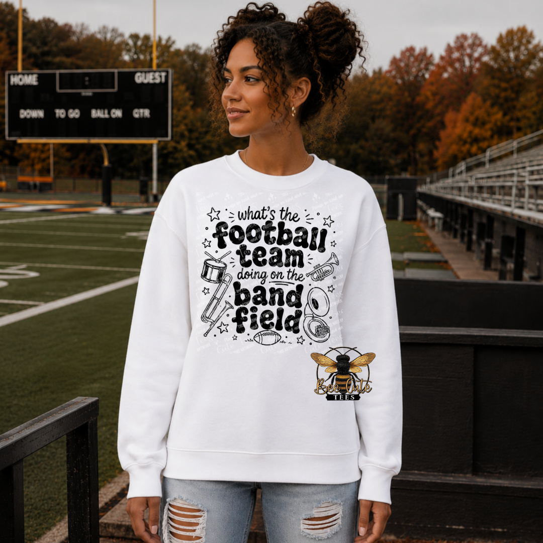 A woman stands on a football field wearing a white crewneck sweatshirt with a black graphic that reads 'what's the football team doing on the band field' along with sports doodles and a bee logo. She is wearing light-wash distressed jeans.