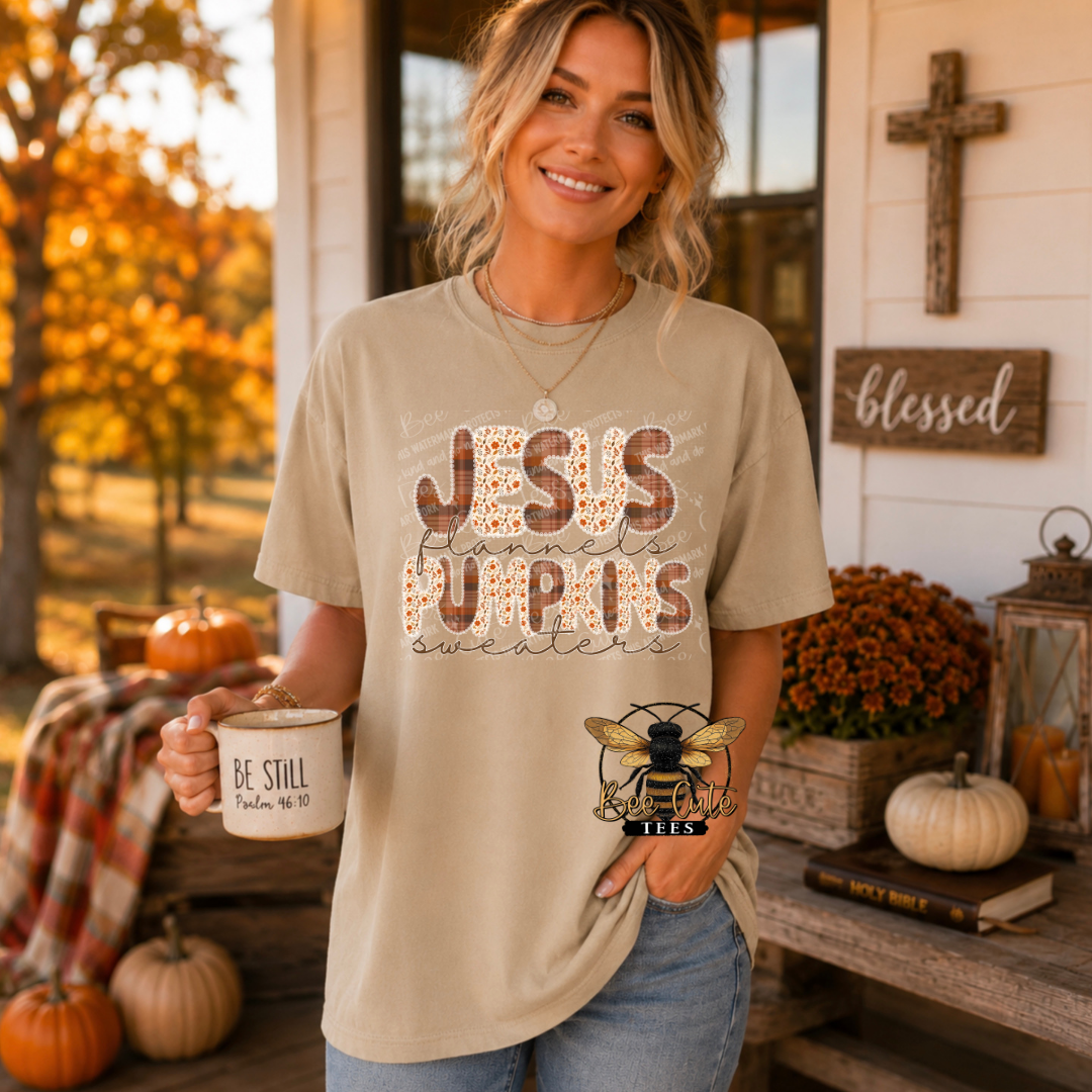 A woman stands on a decorated porch wearing a beige graphic T-shirt that reads 'JESUS PUMPKINS' with a floral pattern and 'sweaters' underneath, holding a mug that says 'BE STILL'.