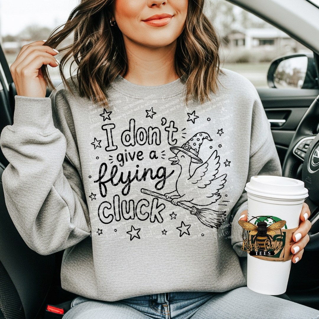A woman sitting in a car wearing a light gray sweatshirt with a black graphic that reads 'I don't give a flying cluck' with a witch, broom, and stars. She is holding a white coffee cup.