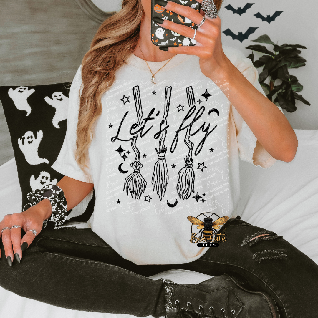 A woman takes a mirror selfie while wearing a white graphic t-shirt with a black 'Let's fly' broomstick design, paired with distressed black jeans and lace-up boots. She is sitting on a bed with a ghost-patterned pillow.