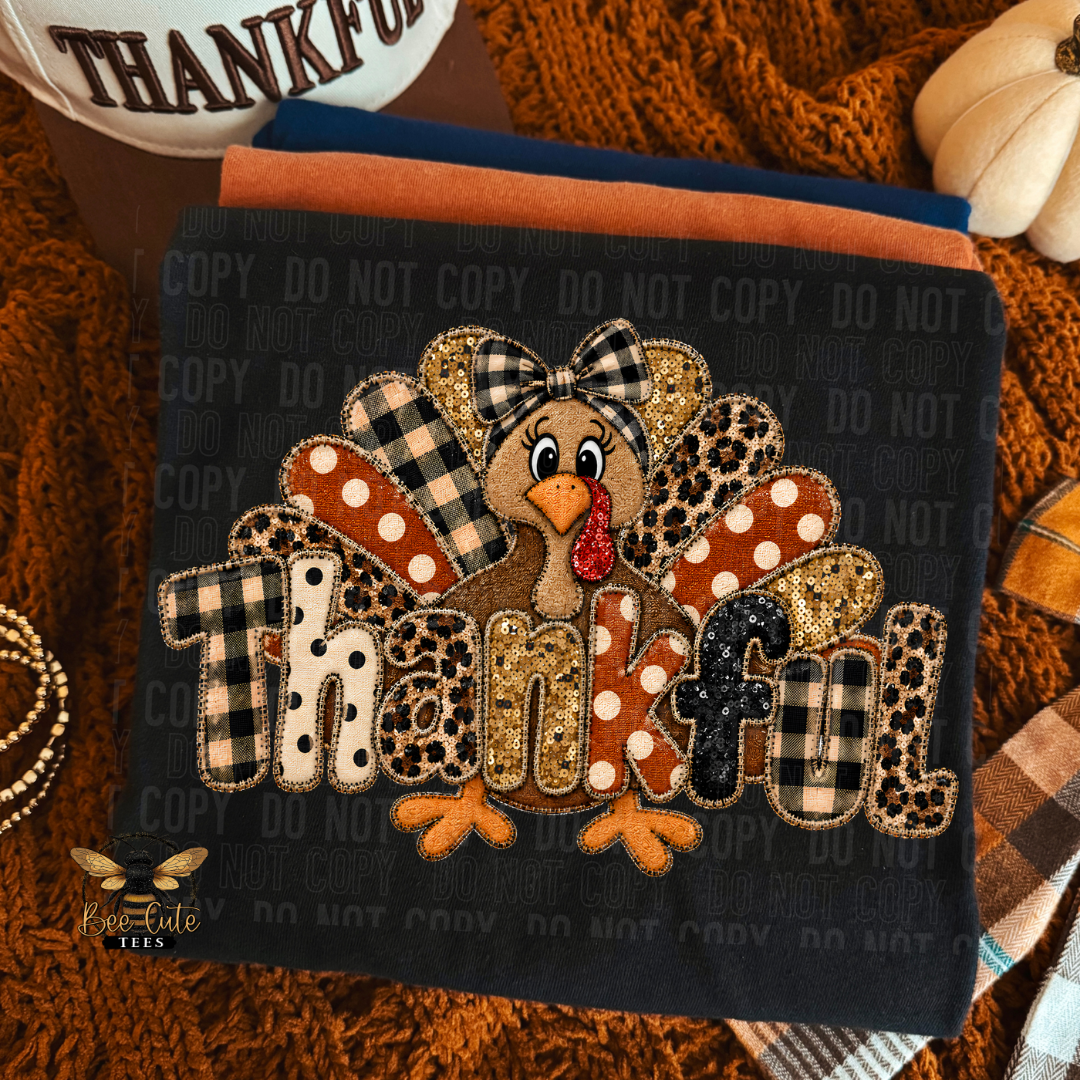 A folded black T-shirt, featuring a detailed  design of a cartoon turkey and the word 'Thankful' in autumn-themed patterned letters. The fabric is styled in a cozy fall setting with a brown knit blanket, a pumpkin, and other folded textiles.