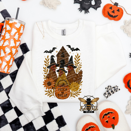 A white long-sleeve sweatshirt with a Halloween-themed graphic print featuring a haunted house, chickens, witch hats, bats, sunflowers, and the words 'TRICK OR TREAT'. The sweatshirt is styled in a flat lay with Halloween props including a ghost-patterned tumbler, black-and-white checkered fabric, plush pumpkins, and white slippers on a white background.