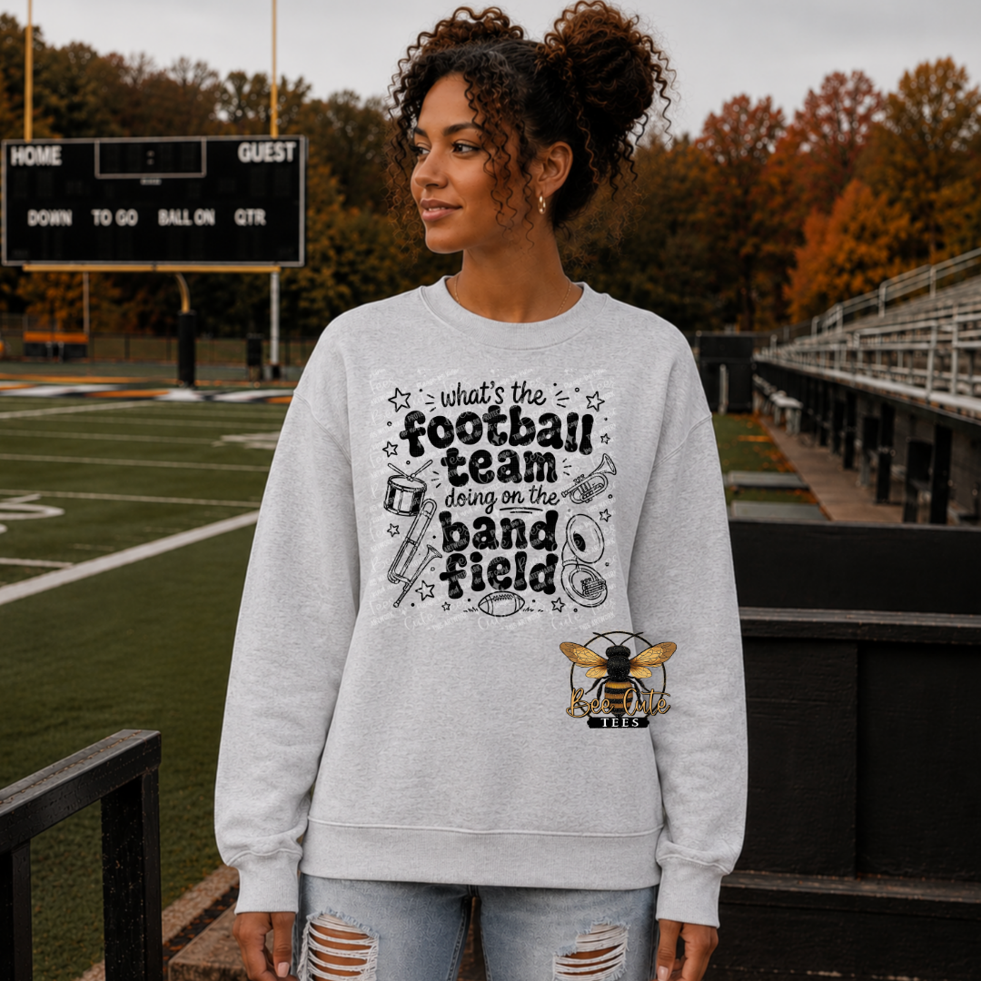 A woman stands on a football field wearing a light gray crewneck sweatshirt with a black and yellow graphic that reads 'what's the football team doing on the band field' along with a bee logo. The background shows the green turf, bleachers, and autumn trees.