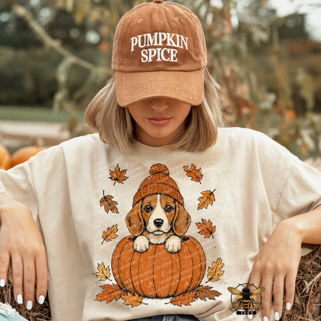 A woman wearing a tan baseball cap that says 'PUMPKIN SPICE' and a cream-colored graphic t-shirt featuring a  puppy sitting in an orange pumpkin surrounded by autumn leaves. She is posed outdoors in a pumpkin patch setting.