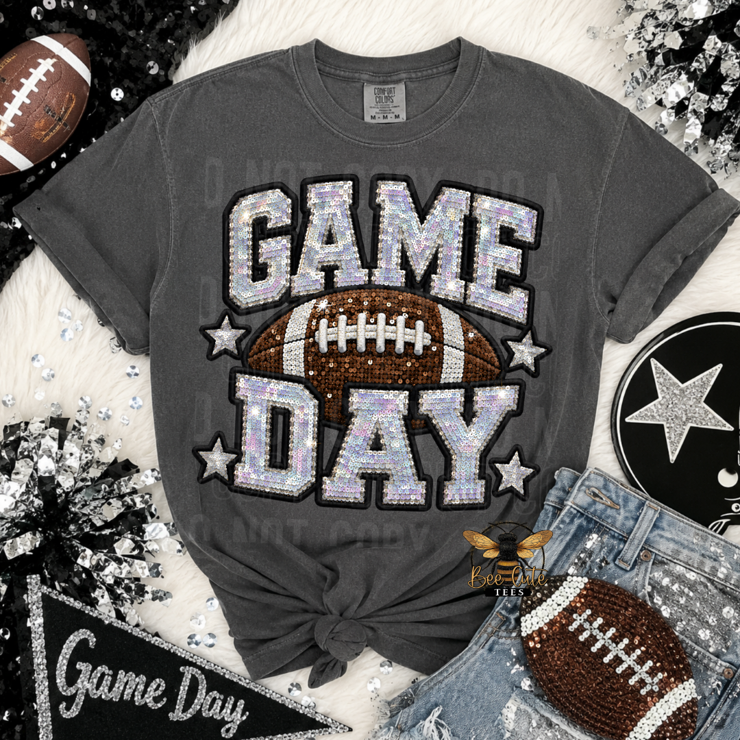 A dark gray graphic t-shirt with 'GAME DAY' in sparkling sequin-style letters and a brown football graphic in the center. The shirt is styled in a flat-lay arrangement with jeans, a rhinestone football, a black star patch, and other football-themed props on a white faux fur background.