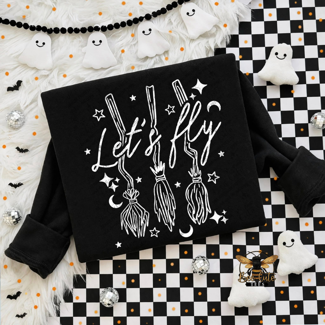 A black long-sleeve sweatshirt laid flat on a Halloween-themed background. The sweatshirt has a white graphic print featuring the phrase 'Let's fly' in script, three broomsticks, stars, and moons.