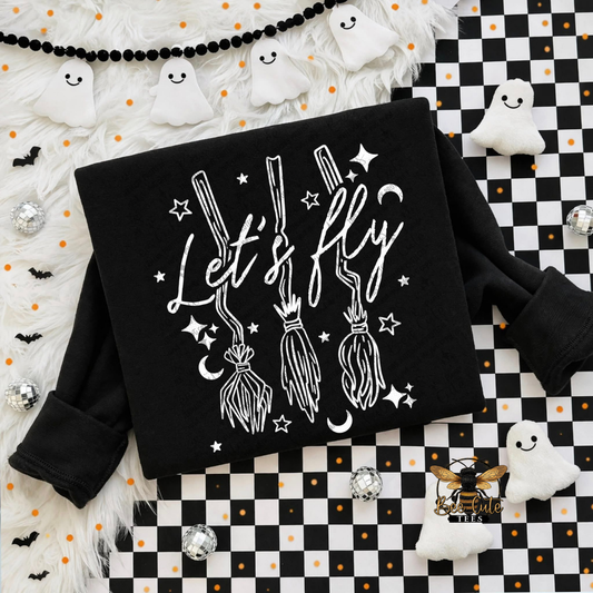 A black long-sleeve sweatshirt laid flat on a Halloween-themed background. The sweatshirt has a white graphic print featuring the phrase 'Let's fly' in script, three broomsticks, stars, and moons.