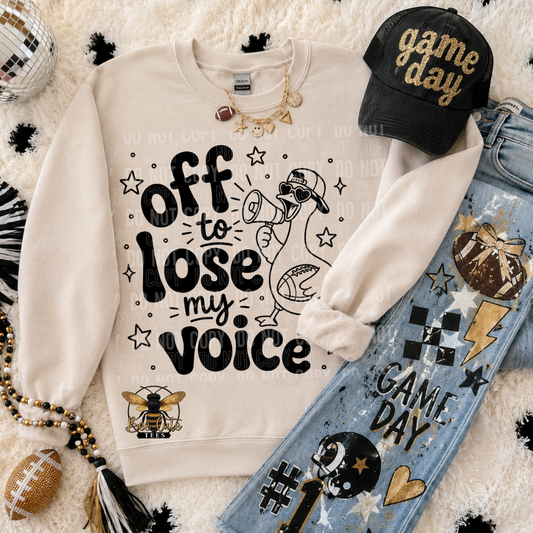 A styled flat-lay featuring a cream sweatshirt with the text 'off to lose my voice' and a duck graphic, arranged with a black 'game day' cap, blue jeans, a football, and accessories on a white faux-fur background.
