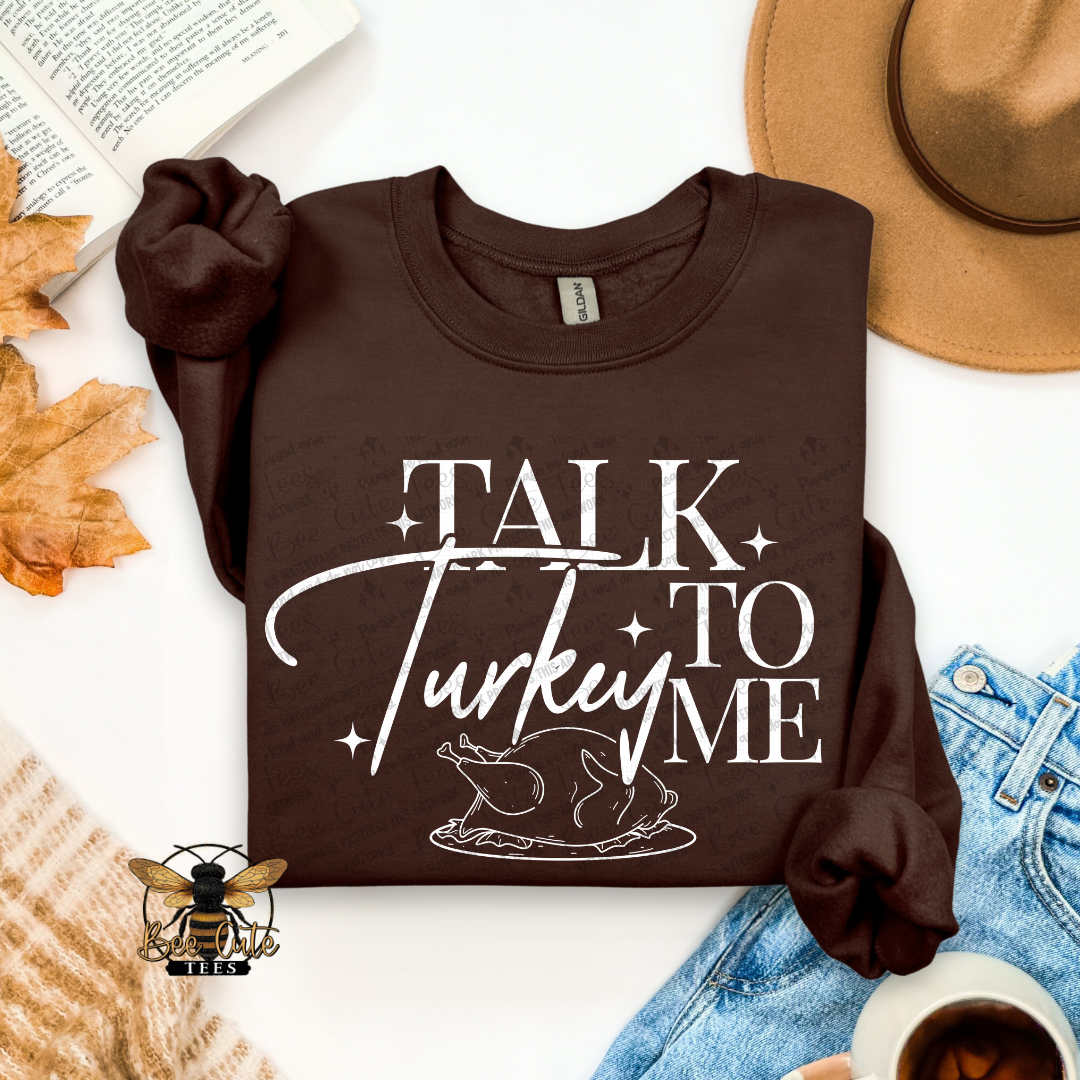 A dark brown long-sleeve t-shirt with a white graphic reading 'TALK Turkey TO ME' and a turkey illustration, styled in a flat lay with a tan hat, blue jeans, a book, autumn leaves, a knit sweater, and a coffee cup on a white background.