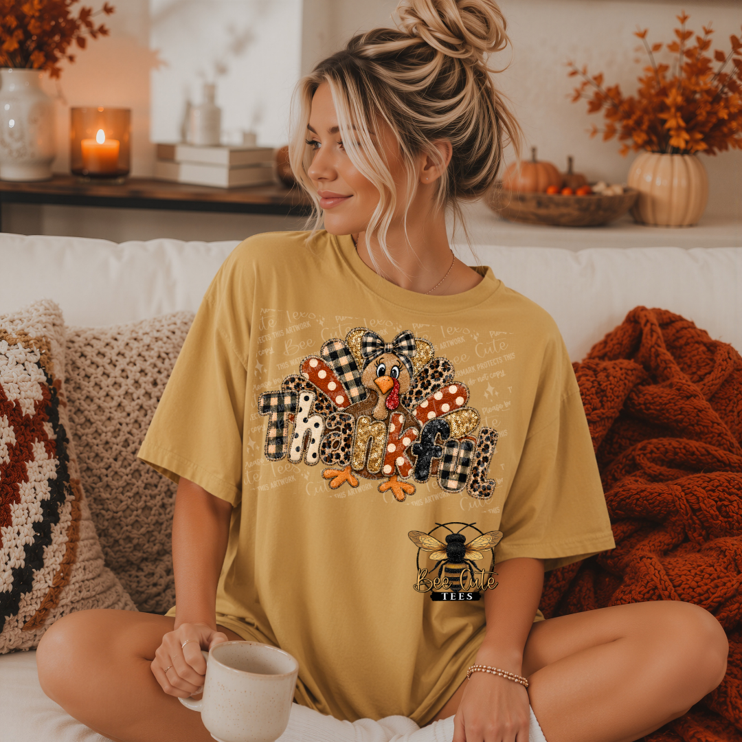 A woman sits cross-legged on a white couch wearing a mustard yellow graphic t-shirt with a turkey. The background features cozy fall decor, including a knitted rust-orange throw blanket, patterned pillows, and a lit candle.