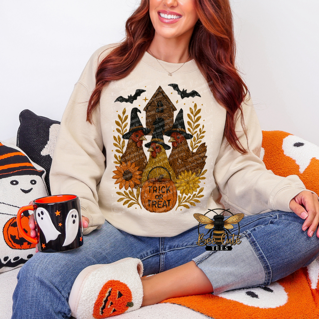 A woman wearing a beige crewneck sweatshirt with a rustic Halloween graphic of three chickens wearing witch hats, surrounded by sunflowers, bats, and the text 'TRICK OR TREAT'. She is sitting on an orange and white blanket with a ghost pillow and holding a black and orange mug.
